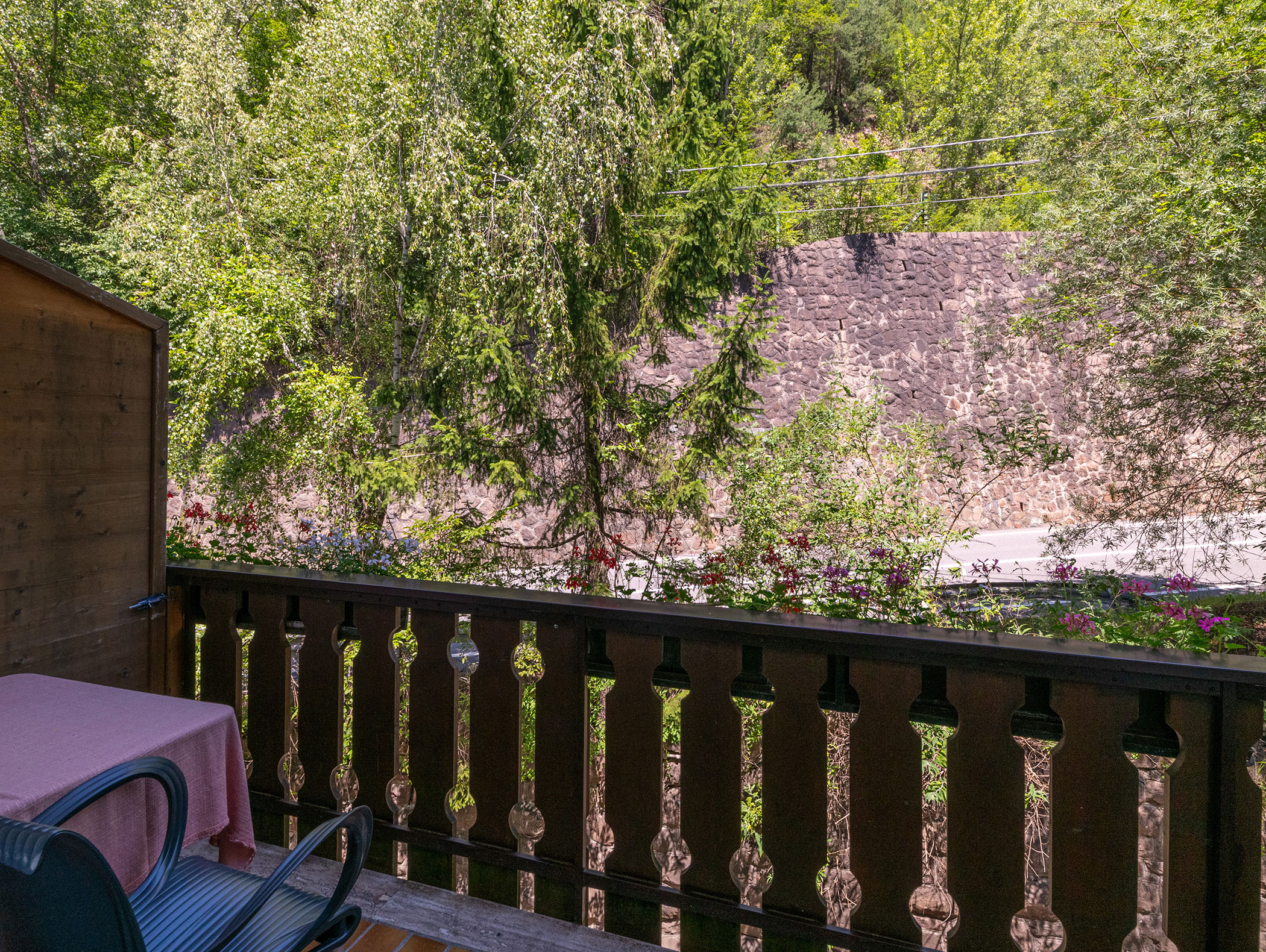 Il balcone, con sedie e tavolo, si affaccia su alberi rigogliosi e, sullo sfondo, su un muro di pietra. - Hotel Zur Sonne