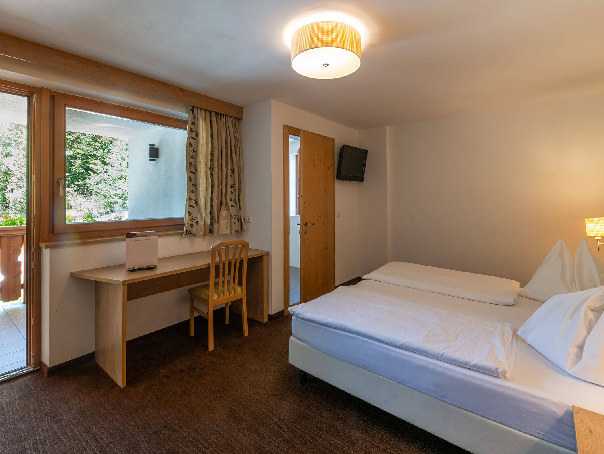 Camera d'albergo con due letti, una scrivania e una sedia, TV appesa alla parete, finestra e porta che dà sul balcone con alberi all'esterno. - Hotel Zur Sonne