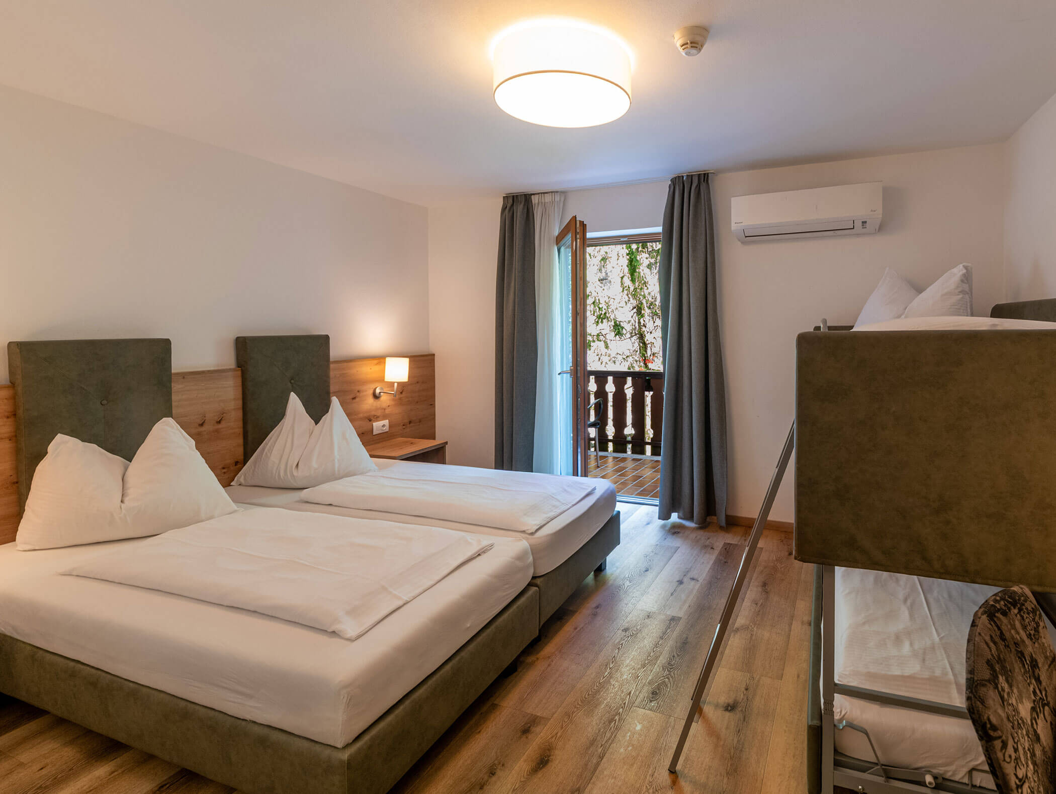Camera d'albergo con due letti singoli, un letto a castello, pavimento in legno e una porta che dà su un balcone con alberi all'esterno. - Hotel Zur Sonne