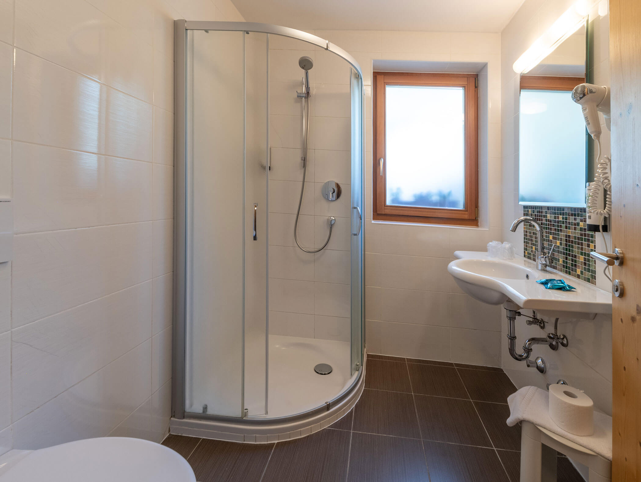 Bagno moderno con box doccia in vetro, pavimento in piastrelle marroni, lavabo bianco e finestra con vetro smerigliato. - Hotel Zur Sonne