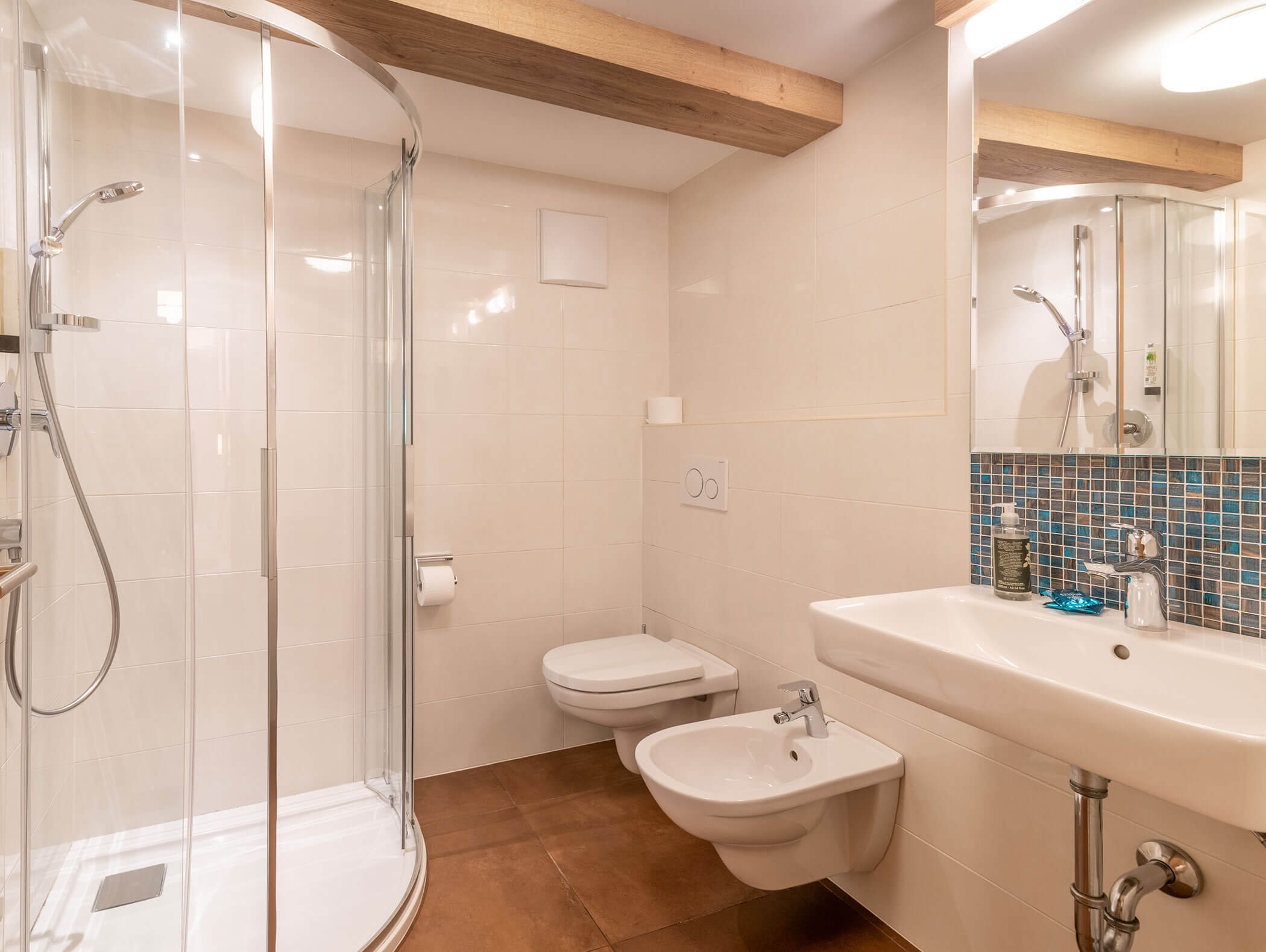 Bagno moderno con box doccia in vetro, WC sospeso, bidet, lavabo e alzatina in piastrelle a mosaico blu. - Hotel Zur Sonne