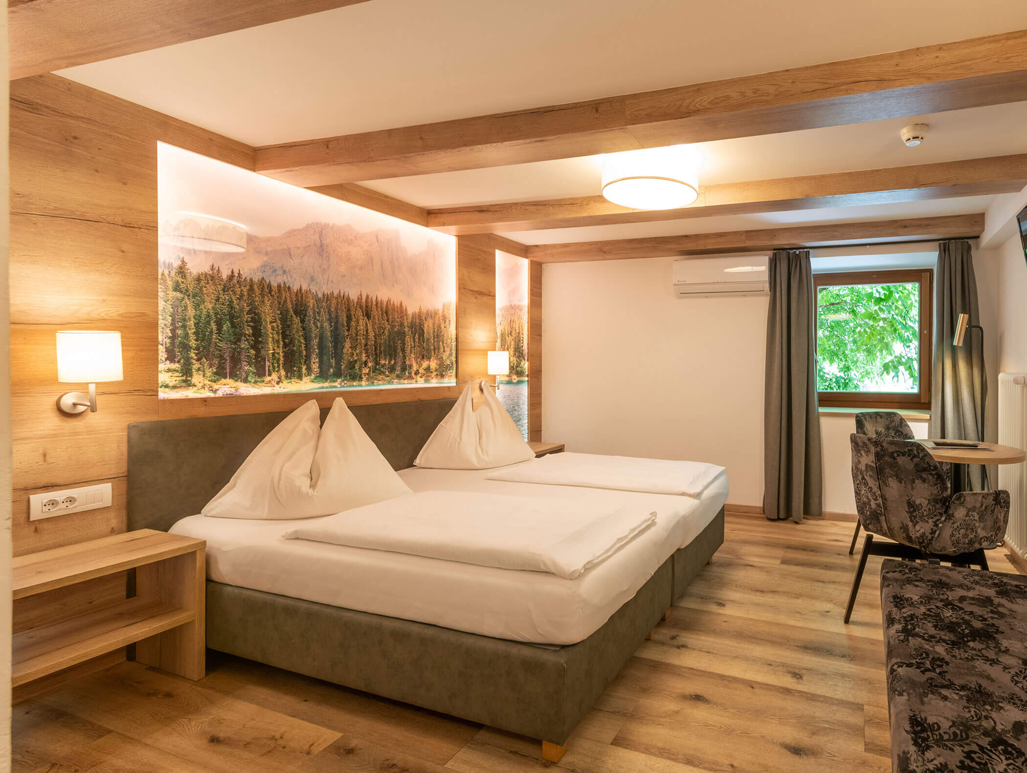 Moderna camera d'albergo con due letti, dettagli in legno, un murale con motivi naturali, una finestra e un tavolino con sedie. - Hotel Zur Sonne