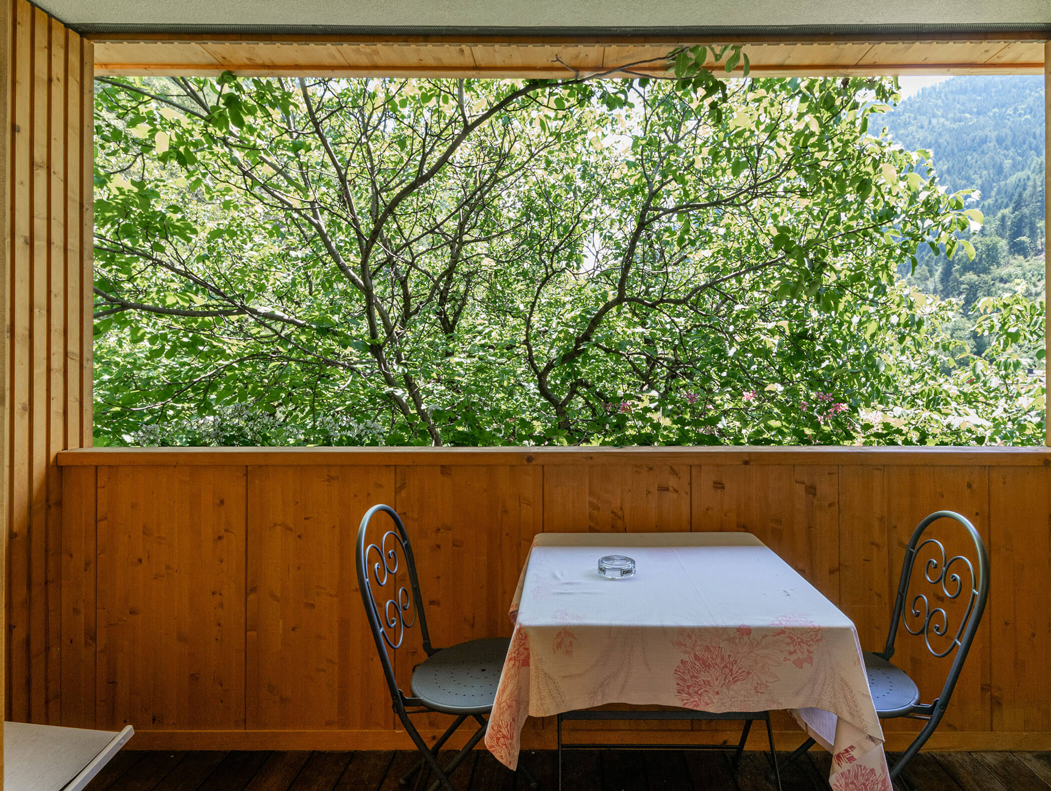 Due sedie e un tavolo con una tovaglia su un balcone in legno, affacciato su alberi verdeggianti e colline in lontananza. - Hotel Zur Sonne