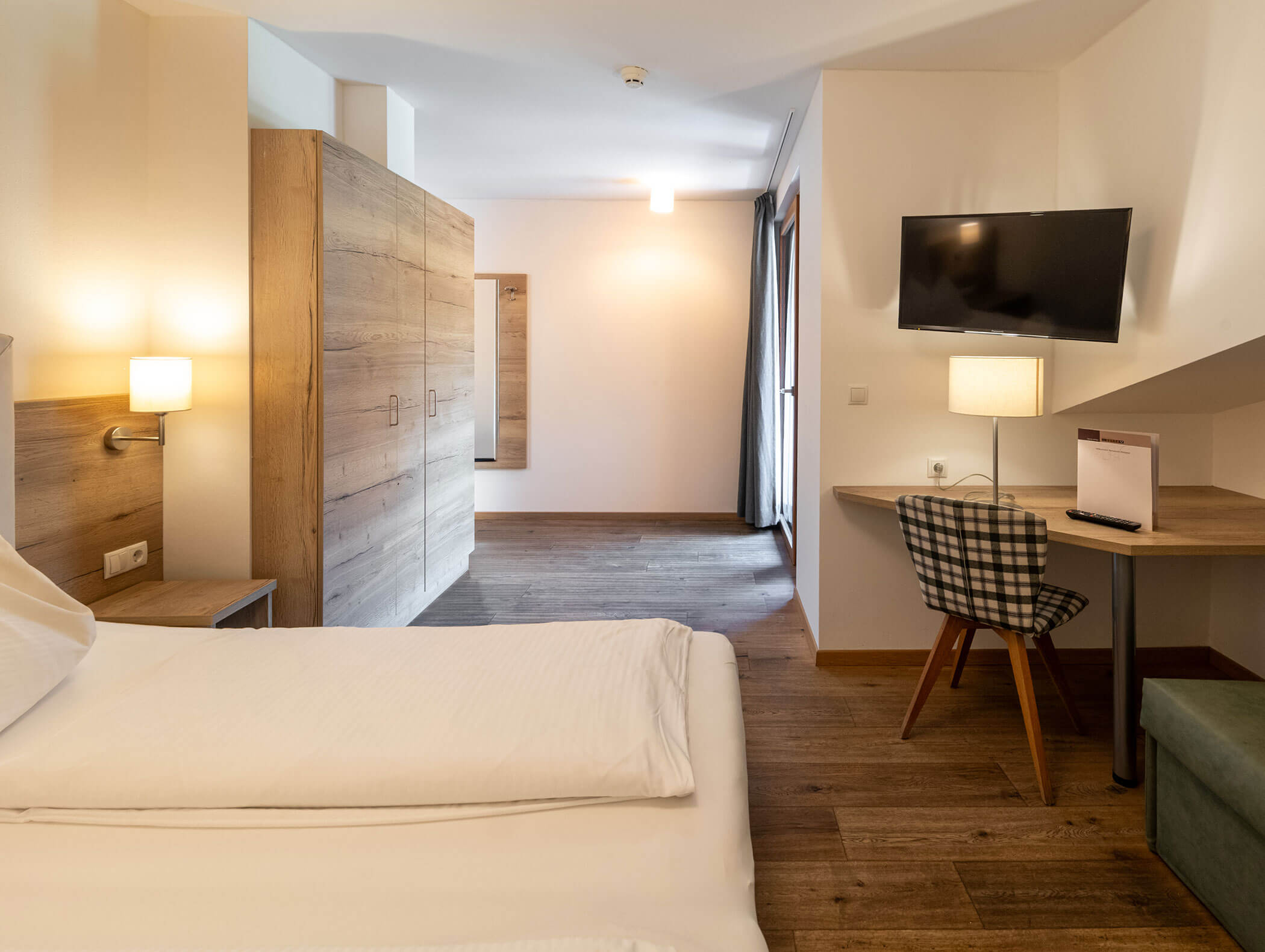 Camera d'albergo luminosa con letto matrimoniale, scrivania, sedia, TV a parete, armadio e pavimento in legno. - Hotel Zur Sonne