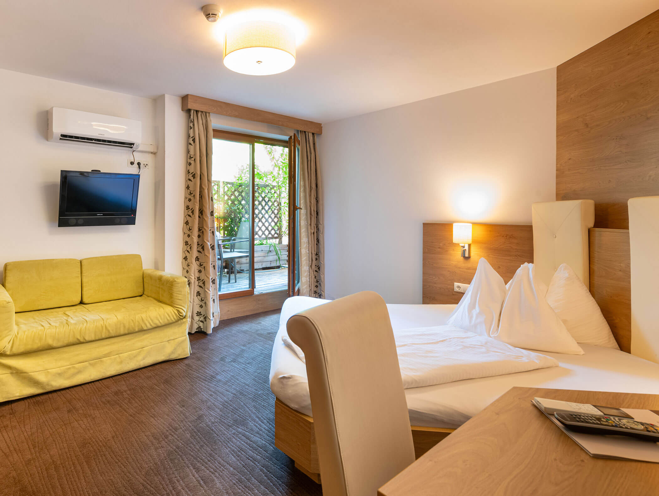 Camera d'albergo luminosa con un letto, un divano giallo, una TV, una scrivania e una porta scorrevole che dà su un patio con posti a sedere all'aperto. - Hotel Zur Sonne