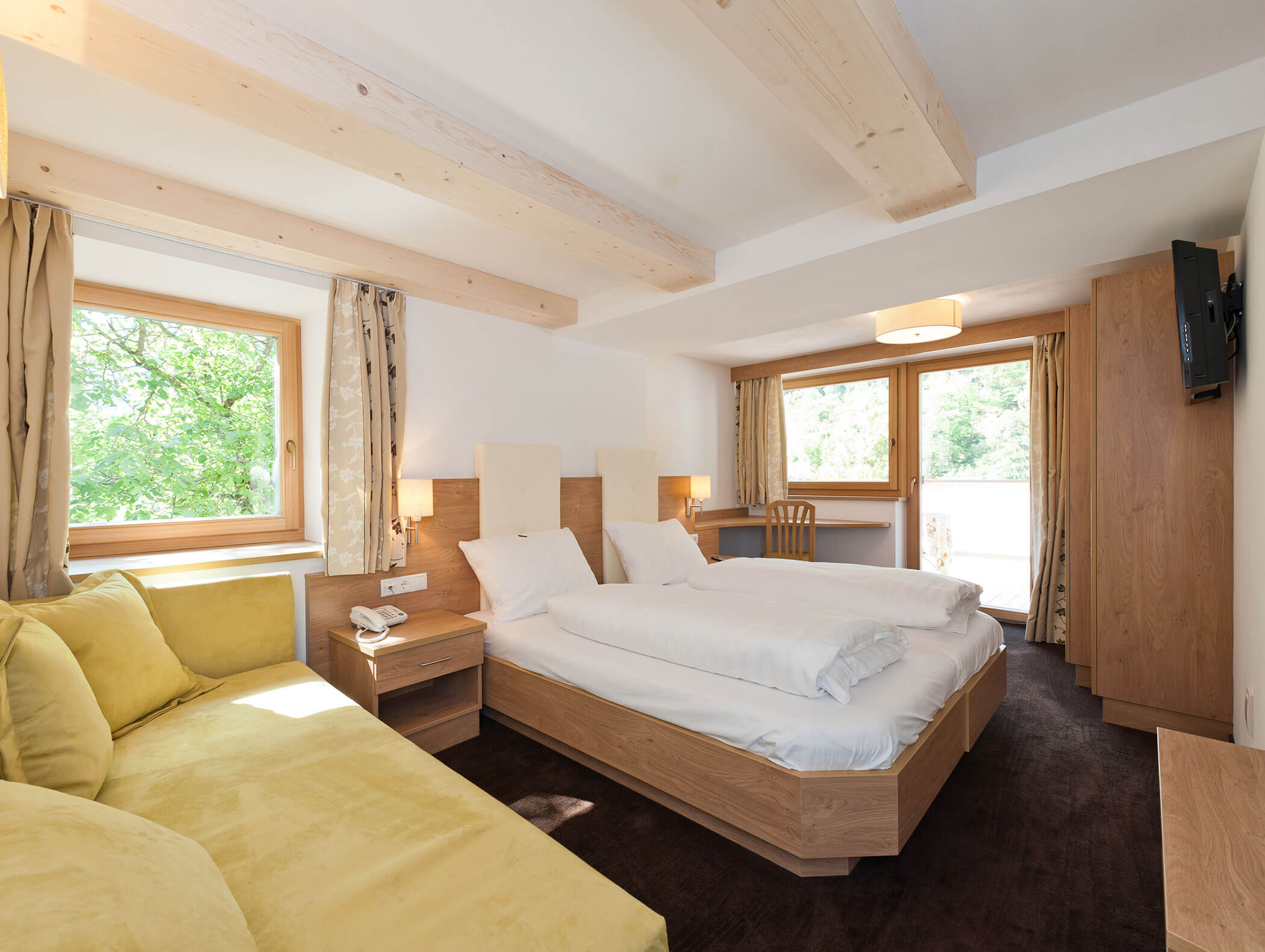 Camera d'albergo luminosa con letto matrimoniale, divano giallo, scrivania, ampie finestre, travi a vista e luce naturale. - Hotel Zur Sonne
